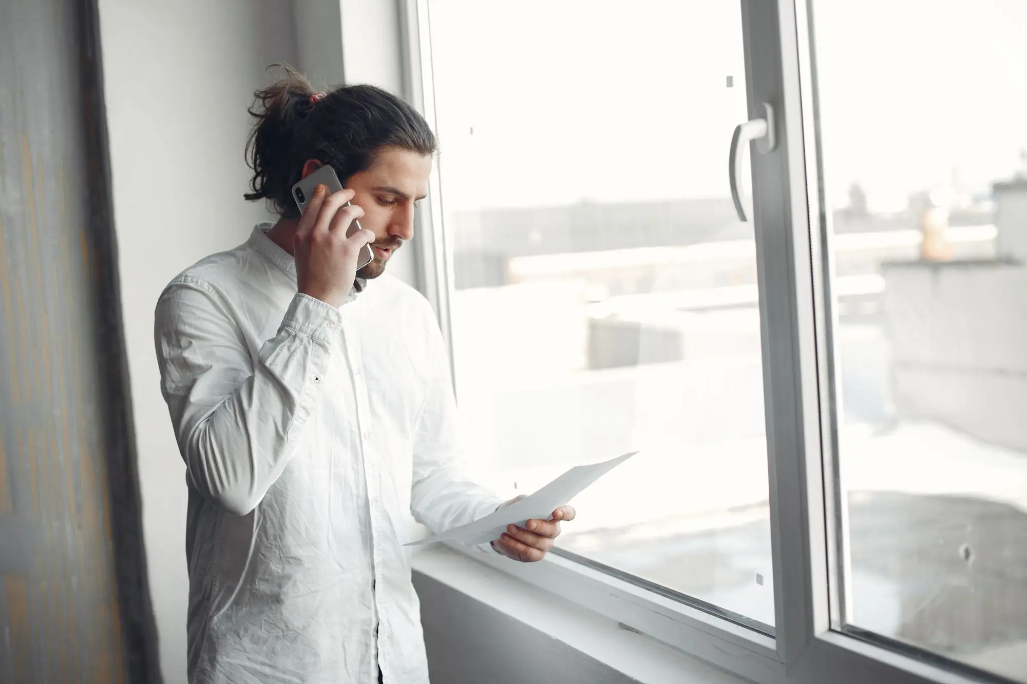Business owner on a phone call by the office window, checking a letter while the caller talks